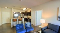 This single-floor residence showcases an open-concept layout that integrates the living and kitchen areas into one cohesive, functional space. The kitchen is outfitted with dark espresso cabinetry, stainless steel appliances including a refrigerator, microwave, and dishwasher, and is anchored by a peninsula-style breakfast bar with seating for multiple guests. Pendant lighting hangs above the counter, adding a warm, inviting glow to the space. The kitchen also features a dedicated sink along the back wall, ample counter space, and open shelving above the upper cabinets for decorative storage. Flowing seamlessly from the kitchen, the living area is finished with hardwood-style flooring that runs throughout, contributing to a clean and modern aesthetic. The living space is well-proportioned, accommodating seating arrangements comfortably. Neutral-toned walls provide a versatile backdrop, while soft ambient lighting from a floor lamp enhances the warmth of the room. A standard interior door near the kitchen provides access to an adjacent room, adding to the functional layout of this well-designed single-level space.