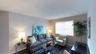 This single-floor space showcases a thoughtfully arranged living area that balances comfort and style. The room is finished with neutral-toned walls that provide a clean, versatile backdrop, complemented by smooth white ceilings that help enhance the sense of openness. Dark hardwood-style flooring anchors the space and contrasts elegantly with the lighter wall tones.

The living area is centered around a plush, dark grey sectional sofa adorned with patterned accent pillows, paired with a matching patterned armchair that adds visual interest without overwhelming the space. A sleek rectangular coffee table sits atop a deep-toned area rug, adding texture and definition to the seating arrangement. Warm ambient lighting from table lamps on either side of the room creates an inviting glow.

A prominent window fitted with white horizontal blinds allows generous natural light to fill the space while maintaining privacy. A vibrant piece of wall art featuring bold blue circular patterns serves as a striking focal point, adding a pop of color to the otherwise neutral palette. The overall ambiance is relaxed yet polished, making it a functional and aesthetically pleasing living environment.