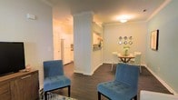 This single-floor residence showcases a thoughtfully designed open-concept layout that promotes a natural flow between the living, dining, and kitchen areas. Architectural columns with crown molding detailing serve as elegant structural dividers, adding visual interest while maintaining the openness of the space.

The living area features rich, dark hardwood flooring that extends throughout, creating a warm and cohesive aesthetic. Tufted blue accent chairs provide stylish seating and a pop of color against the neutral, light gray walls.

The dining area is positioned adjacent to the living space and accommodates a round table with upholstered seating. Decorative diamond-shaped wall mirrors add a contemporary touch above the dining setting.

A half-wall with a pass-through connects to the kitchen, which is finished with white cabinetry and appliances, contributing to a bright and clean appearance. Crown molding runs consistently along the ceilings throughout the space, reinforcing the refined character of the interior. Recessed and flush-mount lighting provides functional illumination across all areas, completing this well-appointed residence.