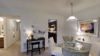This single-floor residence offers a welcoming and functional layout designed to maximize both comfort and practicality. The dining area serves as a central gathering space, featuring a glass-top dining table surrounded by stylishly upholstered seating, all set beneath an elegant pendant light that adds a warm, inviting glow. Soft, neutral-toned carpeting underfoot complements the light-colored walls, creating a bright and airy atmosphere throughout. A charming console table along the wall, accented by a decorative round mirror and artistic wall pieces, adds a personalized touch to the space. Adjacent to the dining area, the kitchen is accessible through an open doorway, showcasing dark cabinetry that provides a rich contrast to the lighter surroundings. The kitchen is equipped with essential appliances and ample cabinetry for storage, making it both practical and visually appealing. A textured ceiling adds subtle depth to the interior, while the overall layout promotes an easy flow between living and dining areas. The space strikes a balance between cozy and functional, making it well-suited for daily living and casual entertaining alike.