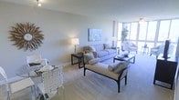 This refined single-floor residence offers a thoughtfully designed open-plan layout that seamlessly blends living and dining areas into one cohesive, light-filled space. The living area is anchored by a plush sofa ensemble in soft, neutral tones, complemented by a chaise bench and accent chairs that create an inviting and well-balanced arrangement. A contemporary abstract artwork adorns the wall above the sofa, adding a touch of color and artistic character to the space. Floor-to-ceiling windows along one wall flood the interior with an abundance of natural light, while offering expansive views of the surrounding cityscape. The lush greenery of an indoor plant beside the windows adds a refreshing organic element to the modern aesthetic. The dining area, positioned adjacent to the living space, features a sleek glass-top table paired with woven accent chairs, ideal for both casual meals and entertaining. Soft, neutral wall tones and plush carpeted flooring run throughout, creating a warm and cohesive ambiance. The overall design strikes a balance between contemporary elegance and comfortable livability, making this a bright, airy, and welcoming space.