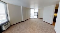 This single-floor space presents a clean, open layout designed to maximize both comfort and functionality. The main living area is finished with wall-to-wall neutral-toned carpet, creating a warm and inviting atmosphere underfoot. Crisp white walls and a smooth white ceiling contribute to a bright, airy feel throughout the room.

A standout feature of this space is the expansive floor-to-ceiling window paired with a sliding glass door, which floods the interior with abundant natural light and provides direct access to an outdoor area. The large glazed opening frames exterior views, adding a sense of openness and connection to the outside environment.

Dark wood baseboards run along the perimeter of the room, offering a subtle contrast against the light walls and flooring. The layout is thoughtfully open, providing flexibility for a variety of furniture arrangements and uses. Structural column detailing adds a minor architectural element to the space without interrupting the overall flow.

The overall ambiance is calm, neutral, and ready for customization, making this a versatile space well-suited to a range of occupants and purposes.
