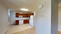 This single-floor unit offers a practical and comfortable layout, thoughtfully organized to make the most of its available space. The kitchen serves as a functional hub, outfitted with rich warm-toned wood cabinetry featuring decorative arched door panels, light countertops, and a full suite of white appliances including an electric range with overhead ventilation, a dishwasher, a refrigerator, and a double basin sink. The tile-style flooring in the kitchen is easy to maintain and complements the cabinetry's warm tones. A full bathroom includes a tiled tub-and-shower combination with a grab bar, a toilet, and a vanity with overhead storage cabinetry — finished with neutral tile flooring for a clean, classic look. The living areas and bedrooms are carpeted in a neutral beige tone, providing a soft underfoot feel throughout. Baseboard heating runs along the walls, ensuring efficient warmth. Multiple double-hung windows flood the living and sleeping areas with natural light and frame lush, green tree-lined views, lending a serene and connected-to-nature quality to the interior. The walls throughout are painted in soft, neutral gray tones, creating a calm and versatile backdrop ready for personalization.
