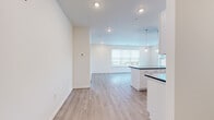 This single-floor unit presents a clean, modern design highlighted by an open-concept layout that seamlessly connects the kitchen and living area. The kitchen is well-appointed with white shaker cabinetry, dark granite countertops, a central island with pendant lighting, and a full suite of stainless steel appliances, including a refrigerator, range, microwave, and dishwasher. Light wood-tone luxury vinyl plank flooring runs throughout the main living areas, complementing the bright white walls and recessed lighting that contribute to a fresh, airy atmosphere. A large window in the living area allows natural light to fill the space while providing views to the exterior. The bedroom features neutral carpet flooring and direct access to a spacious walk-in closet outfitted with wire shelving and hanging rods, offering generous storage. An additional room with wood-tone flooring and a wide window provides versatile living space. The bathroom is finished with white cabinetry, a white countertop, and matching light wood-tone flooring, maintaining the cohesive, contemporary aesthetic carried throughout the unit.