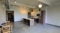 This single-floor space in Dallas, TX presents a clean, modern aesthetic defined by an open-concept layout that allows for seamless flow between living and kitchen areas. Polished concrete floors extend throughout, lending an industrial yet refined character to the interior. The soaring ceilings amplify the sense of openness, while a combination of track lighting and a contemporary globe pendant light ensures the space is well-illuminated from above.

The kitchen is a standout feature, outfitted with modern two-tone cabinetry in a warm wood-grain finish complemented by crisp upper units. A central island with a light quartz countertop provides additional workspace and anchors the kitchen within the open floor plan. Stainless steel appliances, including a refrigerator, microwave, and range, complete the functional and stylish cooking area. A dedicated sink is neatly integrated into the countertop run, and subtle greenery accents atop the cabinetry add a touch of life to the space.

The open living area offers generous room for flexible furniture arrangements, with neutral white walls serving as a versatile backdrop. The overall ambiance is contemporary and minimalist, making this Dallas space well-suited for those who appreciate clean lines, industrial design elements, and modern urban living.