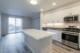 This single-floor residence presents a clean, contemporary design highlighted by light wood-look flooring that runs consistently throughout the space, creating a cohesive and airy feel. The open-concept kitchen and living area serves as the heart of the unit, anchored by a striking white quartz island with subtle veining that provides both additional workspace and a visual focal point. The kitchen is well-appointed with flat-front white cabinetry extending to the ceiling for maximum storage, dark countertops, and a full suite of stainless steel appliances including a gas range, built-in microwave, dishwasher, and a side-by-side refrigerator with an in-door water and ice dispenser. Under-cabinet lighting adds both functionality and ambiance. The living area extends naturally from the kitchen and opens to a sliding glass door, allowing natural light to flow in and providing outdoor access. The bedroom features a natural light source via a window with blinds and connects to a dedicated walk-in closet with built-in shelving and hanging space. The bathroom is finished in a clean, neutral palette with a tiled shower-tub combination, white vanity with dark hardware, dark countertop, and a large mirror. A well-lit hallway connects the rooms seamlessly.