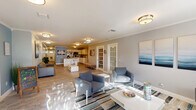 Located at 1601 Northwest 13th Street in Boca Raton, FL, this single-floor space offers a well-designed open-concept layout that seamlessly integrates a fully equipped kitchen with a generous living area. The interior is finished with wood-look tile flooring throughout, crown molding, and a soothing coastal color palette of soft grays and muted blues that creates a calm, welcoming ambiance.

The kitchen is outfitted with white shaker-style cabinetry, a stainless steel refrigerator, microwave, and a deep single-basin sink. A central island with a white quartz countertop and bar stool seating serves as both a functional workspace and a casual gathering point. Ample cabinet storage lines the walls, providing excellent organization.

The living area is generously proportioned, featuring multiple seating arrangements including blue upholstered chairs, a leather sectional sofa, and white accent tables atop a striped area rug. Natural light streams in through windows and is complemented by flush-mount ceiling fixtures. Coastal-themed artwork adorns the walls, enhancing the space's relaxed aesthetic.

French glass-paned doors lead to adjacent rooms, adding architectural character and allowing light to flow freely. The open floor plan and cohesive design make this Boca Raton space both functional and visually inviting.
