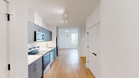 Located on the sixth floor at 303 E Main St in Bound Brook, NJ, this efficiently designed unit offers a clean, contemporary aesthetic throughout its single-level layout. The open-plan kitchen and living area forms the heart of the space, where light wood-tone flooring and crisp white walls create a bright, modern atmosphere. The kitchen is outfitted with slate-blue cabinetry—both upper and lower—paired with white countertops and a full suite of stainless steel appliances, including a refrigerator, electric range, dishwasher, and over-the-range microwave. Rail track lighting runs along the kitchen corridor, adding both functionality and a modern design touch. The living area is bathed in natural light courtesy of large, floor-to-ceiling windows that frame elevated views of downtown Bound Brook and the surrounding streetscape. A wall-mounted HVAC unit is positioned beneath the windows for year-round climate control. The bathroom features large-format white tile flooring, a frameless glass walk-in shower with matching tile surround, and a clean, minimalist finish throughout. Closet space is provided via double-door closets visible from the main living area. The overall design is cohesive and contemporary, emphasizing clean lines, neutral tones, and efficient use of space.