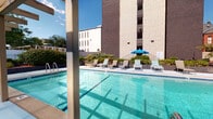 This outdoor pool area offers a well-appointed recreational retreat designed for relaxation and leisure. The centerpiece is a generously sized swimming pool with clear turquoise water, accessible via stainless steel entry rails and framed by a wide concrete sundeck. Along one side of the deck, a row of lounge chairs and a shaded umbrella provide ample space for sunbathing, while a wooden pergola structure on the opposite side offers a partially shaded lounging zone furnished with reclining chairs. A distinctive decorative breeze-block wall runs alongside the pergola area, featuring a geometric, lattice-like pattern that adds architectural character while providing partial privacy screening. The pool area is surrounded by manicured shrubs and greenery, with mature trees visible beyond the perimeter, creating a pleasant natural buffer. The backdrop includes the multi-story brick building the pool serves, along with views of the surrounding urban neighborhood under open blue skies. The combination of the pergola, decorative masonry wall, and open sundeck creates a versatile outdoor environment suited to both active recreation and quiet relaxation.