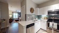 This single-floor residence offers a thoughtfully designed layout that balances style and functionality throughout. The kitchen is a standout feature, equipped with stainless steel appliances including a range, microwave, dishwasher, and refrigerator. Crisp white cabinetry, light quartz countertops, and a classic grey subway tile backsplash lend a clean, contemporary aesthetic, while the peninsula provides additional prep space and opens directly to the living area. Dark hardwood-style flooring runs through the main living spaces, anchoring the open-plan layout. The living and dining area is defined by a bold blue accent wall and features a dedicated dining nook beneath a pendant light, offering a comfortable and visually dynamic gathering space with natural light from surrounding windows. The bedroom features a tray ceiling with a ceiling fan, plush carpet flooring, and a warm yellow accent wall that adds a vibrant, energetic character to the room. Large windows invite generous natural light into this restful retreat. The en-suite bathroom includes a dual-sink vanity with white cabinetry, a large mirror, tile flooring, and direct access to a spacious walk-in closet complete with multi-rod shelving and ample storage space, providing a practical and organized solution for everyday living.