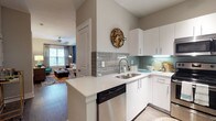 This single-floor residence offers a thoughtfully designed layout that balances style and functionality throughout. The kitchen is a standout feature, equipped with stainless steel appliances including a range, microwave, dishwasher, and refrigerator. Crisp white cabinetry, light quartz countertops, and a classic grey subway tile backsplash lend a clean, contemporary aesthetic, while the peninsula provides additional prep space and opens directly to the living area. Dark hardwood-style flooring runs through the main living spaces, anchoring the open-plan layout. The living and dining area is defined by a bold blue accent wall and features a dedicated dining nook beneath a pendant light, offering a comfortable and visually dynamic gathering space with natural light from surrounding windows. The bedroom features a tray ceiling with a ceiling fan, plush carpet flooring, and a warm yellow accent wall that adds a vibrant, energetic character to the room. Large windows invite generous natural light into this restful retreat. The en-suite bathroom includes a dual-sink vanity with white cabinetry, a large mirror, tile flooring, and direct access to a spacious walk-in closet complete with multi-rod shelving and ample storage space, providing a practical and organized solution for everyday living.