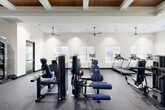Located in Glendale, Arizona, this well-appointed fitness center offers a generously sized, single-floor exercise space designed to support a comprehensive workout experience. The room's most striking architectural feature is its coffered ceiling with rich wood inlays, recessed lighting, and multiple ceiling fans that ensure comfortable airflow throughout the space. Crisp white walls and large, blinds-fitted windows allow natural light to fill the room while maintaining privacy, creating a bright and energizing atmosphere. The durable speckled rubber flooring provides both safety and comfort underfoot, suitable for high-impact activity. The space is fully equipped for a well-rounded fitness routine, with a dedicated cardio zone featuring multiple Spirit treadmills and an elliptical trainer, alongside a strength training area outfitted with a power rack, cable machines, multi-station weight machines, a dumbbell rack, and adjustable benches. A floor-to-ceiling mirror along one wall enhances the sense of openness and allows users to monitor their form. The layout is thoughtfully organized to provide ample room between equipment, ensuring a safe and comfortable environment. Wide double doors offer easy access, completing a fitness facility that is both functional and visually refined.