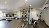 Located at 1598 Crestview Dr in Nashville, Tennessee, this fitness center offers a well-appointed, single-floor exercise space designed to support a comprehensive workout routine. The room features neutral gray walls accented by bold yellow horizontal stripes, crown molding, and a textured ceiling with recessed lighting that keeps the space bright and inviting throughout the day. Multiple windows allow natural light to filter in, while a set of glass French doors provides an additional entry point and further enhances the sense of openness. The flooring consists of durable dark rubberized material suited for high-traffic athletic use. Cardio equipment includes multiple treadmills and elliptical machines positioned along the perimeter, while a multi-station cable strength system serves as the centerpiece of the room, accommodating a variety of resistance training exercises. A rack of medicine balls, a stability ball, and wall-mounted workout reference charts round out the available fitness resources. A large wall mirror spans one side of the room, enhancing the sense of space and allowing users to monitor their form during exercise. A wall-mounted air conditioning unit and ventilation panel ensure climate comfort. Safety features including a fire extinguisher and a sanitation station are also present, reflecting a commitment to a clean and secure workout environment.