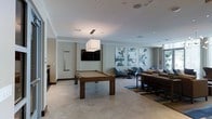 This single-floor amenity lounge is designed to offer a comfortable and engaging communal experience. The space is anchored by a full-sized wooden pool table positioned beneath a sleek square pendant light, creating a natural focal point for recreation. A wall-mounted flat-screen television is installed nearby, adding to the entertainment options available.

Adjacent to the game area, a relaxed lounge zone features contemporary blue accent chairs and low-profile seating arrangements set atop a patterned area rug, encouraging casual conversation and socializing. A long communal table with bench seating extends along one side of the room, providing additional gathering space.

The walls are adorned with a series of large-scale abstract art panels in soft blue and neutral tones, contributing to the modern, curated aesthetic of the space. Light tile flooring runs throughout, complementing the neutral, warm color palette of the walls and furnishings. Recessed lighting and a pendant fixture provide layered illumination, while a window along one wall allows natural light to filter into the room, enhancing the overall bright and welcoming ambiance.