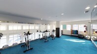 This fitness center offers a clean, modern environment designed to support a variety of workout routines. The space is finished with a striking teal carpet that covers the entire floor, adding a vibrant and energetic atmosphere to the room. A row of spinning bikes is positioned along the perimeter, facing a continuous stretch of large windows fitted with roller shades that allow for adjustable natural light. The bright, white walls and recessed ceiling lighting create a well-lit and open feel throughout. Structural columns are integrated into the layout without disrupting the flow of the space. On the opposite side of the room, a colorful dumbbell rack provides a range of free weight options, making the area suitable for both cardio and strength training. Additional equipment and storage, including a dark storage unit, are thoughtfully placed to maximize usable floor space. The overall design is functional and inviting, with an emphasis on natural light, spaciousness, and accessibility — making it a well-rounded facility for daily exercise needs.