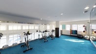 This fitness center offers a clean, modern environment designed to support a variety of workout routines. The space is finished with a striking teal carpet that covers the entire floor, adding a vibrant and energetic atmosphere to the room. A row of spinning bikes is positioned along the perimeter, facing a continuous stretch of large windows fitted with roller shades that allow for adjustable natural light. The bright, white walls and recessed ceiling lighting create a well-lit and open feel throughout. Structural columns are integrated into the layout without disrupting the flow of the space. On the opposite side of the room, a colorful dumbbell rack provides a range of free weight options, making the area suitable for both cardio and strength training. Additional equipment and storage, including a dark storage unit, are thoughtfully placed to maximize usable floor space. The overall design is functional and inviting, with an emphasis on natural light, spaciousness, and accessibility — making it a well-rounded facility for daily exercise needs.
