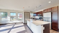 This well-appointed single-floor unit offers a thoughtfully designed layout with a clean, contemporary aesthetic throughout. The heart of the home is an open-concept kitchen featuring rich dark-wood cabinetry extending to the ceiling, a large central island with a bright quartz countertop and integrated sink, and a full suite of stainless steel appliances including a refrigerator, range, and microwave. Pendant lighting overhead and light hardwood-style flooring tie the kitchen seamlessly into the adjoining living area, which is bathed in natural light from an expansive bank of large windows offering neighborhood views. Additional rooms feature plush carpeting, crisp white walls, dark wood trim, and generous windows that bring in ample daylight. The bathroom is well-equipped with rich dark wood vanity cabinetry, stone-look countertops, and a full tub-shower combination, all laid out in a practical, elongated floor plan. Throughout the unit, two-panel interior doors with dark wood trim frames add a consistent design element, while the neutral color palette creates a bright and versatile atmosphere. The overall layout, as visible in the dollhouse view, is efficiently organized and offers a comfortable sense of space and flow.