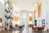This single-floor space makes an immediate impression with its dramatic vaulted ceilings that rise to exceptional heights, anchored by oversized windows trimmed in warm golden yellow that flood the interior with natural light. The open-concept layout is unified throughout by rich herringbone-patterned wood flooring, lending visual continuity across distinct functional zones.

The main lounge area centers around a statement fireplace set within a chevron-patterned wood accent wall, above which a mounted flat-screen television is positioned. The seating arrangement combines eclectic mid-century modern furnishings — including patterned accent chairs, upholstered sofas, and varied side tables — atop a textured area rug. Decorative wall art, metallic sculptural plates, and geometric wood panels add curated character throughout.

A dedicated dining zone features round white tables paired with retro-style burnt orange swivel chairs, contributing to the space's bold, contemporary palette. Pendant light fixtures with gold-toned detailing hang from the soaring ceiling, reinforcing the warm color scheme.

The kitchen is outfitted with vivid blue shaker-style cabinetry, granite countertops, and stainless steel appliances including a refrigerator, range, and built-in microwave, all set against the same herringbone flooring. The overall ambiance is vibrant, energetic, and thoughtfully designed.