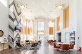 This single-floor space makes an immediate impression with its dramatic vaulted ceilings that rise to exceptional heights, anchored by oversized windows trimmed in warm golden yellow that flood the interior with natural light. The open-concept layout is unified throughout by rich herringbone-patterned wood flooring, lending visual continuity across distinct functional zones.

The main lounge area centers around a statement fireplace set within a chevron-patterned wood accent wall, above which a mounted flat-screen television is positioned. The seating arrangement combines eclectic mid-century modern furnishings — including patterned accent chairs, upholstered sofas, and varied side tables — atop a textured area rug. Decorative wall art, metallic sculptural plates, and geometric wood panels add curated character throughout.

A dedicated dining zone features round white tables paired with retro-style burnt orange swivel chairs, contributing to the space's bold, contemporary palette. Pendant light fixtures with gold-toned detailing hang from the soaring ceiling, reinforcing the warm color scheme.

The kitchen is outfitted with vivid blue shaker-style cabinetry, granite countertops, and stainless steel appliances including a refrigerator, range, and built-in microwave, all set against the same herringbone flooring. The overall ambiance is vibrant, energetic, and thoughtfully designed.