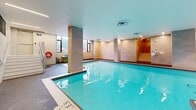 Located at 35 David Salomon Drive in Toronto's M3C neighbourhood, this indoor aquatic facility offers a generously proportioned, single-level pool space designed with both function and comfort in mind. The centrepiece is a large indoor swimming pool featuring clearly marked depth zones, including a designated deep area along one side. The pool is framed by a non-slip aggregate deck surface that provides ample walkway space around the perimeter.

The interior is finished with large-format tile cladding on the walls and structural columns, lending a clean and contemporary aesthetic throughout. A row of windows along one wall allows natural light to filter in, brightening the space while maintaining privacy through frosted lower panes. Recessed and linear ceiling lighting supplements the natural illumination, ensuring consistent brightness across the facility.

A staircase with metal railings provides access to an elevated area at one end of the pool, while accessible entry features such as handrails are integrated along the pool's edge. Safety equipment, including life rings and emergency tools, is strategically positioned around the space. The facility also includes designated areas for pool equipment storage. The smooth white ceiling and neutral-toned finishes throughout create a calm, professional environment well-suited for lap swimming and aquatic programming.