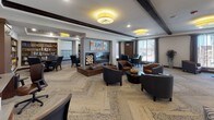 This thoughtfully designed single-floor communal space offers a warm and sophisticated atmosphere through a seamless blend of contemporary and transitional design elements. Striking dark wood ceiling beams contrast beautifully against crisp white ceilings, while distinctive patterned carpeting in a herringbone-inspired layout adds visual texture throughout the open floor plan.

The space is organized into several functional zones. A central lounge area anchors the room with a sleek, modern fireplace flanked by rich leather and upholstered club chairs arranged for comfortable conversation. A dedicated library nook features built-in wooden shelving stocked with books and decorative accents, accompanied by high-back armchairs ideal for quiet reading. A formal dining or meeting area includes a dark wood table surrounded by tufted leather chairs in a sophisticated teal finish.

A separate workstation area provides a wall-mounted flat-screen television and computer desks with task seating, catering to productivity needs. Large windows dressed with sheer curtains allow generous natural light to filter throughout, enhancing the open and airy feel of the space. Stylish drum pendant light fixtures with a warm yellow glow add a refined finishing touch. The overall ambiance is inviting and well-appointed, balancing functionality with upscale residential comfort.