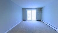 This single-floor unit offers a clean, open living area finished with wall-to-wall carpeting in a neutral tone, providing a comfortable and versatile space. The room is painted in a soft light gray, contributing to a calm and airy atmosphere. A sliding glass door serves as a central architectural feature, drawing in natural light and providing direct access to a private balcony, where views of the surrounding exterior are visible. Vertical blinds accompany the sliding door, offering flexible light control. Climate comfort is addressed through a combination of a wall-mounted air conditioning unit positioned on the exterior wall and baseboard heating units running along the perimeter of the room, ensuring year-round temperature regulation. The white baseboard trim contrasts cleanly against the light-colored walls, adding a crisp, finished look. The space is straightforward in layout, well-lit by natural light from the balcony door, and move-in ready, making it a functional and practical living environment.