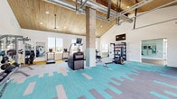 Located at 3229 W Montague Ave in North Charleston, SC, this single-floor fitness center offers a generously sized, well-appointed space designed to accommodate a wide range of workout routines. The facility is divided into two distinct zones, each with its own character and purpose.

The main gym area features soaring vaulted ceilings clad in warm wood paneling, exposed industrial ductwork, and recessed lighting that creates a bright, energizing atmosphere. Striking brick columns add an architectural accent throughout. The floor is covered in a colorful patchwork carpet in teal, gray, and gold tones. This zone is equipped with multiple treadmills, rowing machines, cable machines, a dumbbell rack, a punching bag, and a wall-mounted flat-screen TV. Large glass garage doors open toward an outdoor pool area, flooding the space with natural light and providing a seamless indoor-outdoor connection.

The secondary studio area offers a quieter, more intimate setting with smooth hardwood flooring, soft sage-green walls, and its own vaulted wood ceiling with ceiling fans. Floor-to-ceiling mirrors line one wall, and the room accommodates spin bikes, stability balls, balance trainers, and other functional fitness accessories — making it ideal for cycling classes or focused individual training.