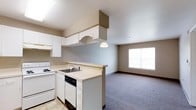 This single-floor space offers a practical and well-organized open-concept layout that connects the kitchen and living area in a cohesive, functional design. The kitchen is equipped with white cabinetry, including overhead and base cabinets that provide ample storage. A built-in overhead cabinet structure above the counter adds a distinctive architectural element to the space. The kitchen features a white electric range, a dishwasher, a stainless steel sink, and light-toned countertops that run along the perimeter and extend into a peninsula, creating a natural division between the kitchen and the adjacent living area. A pendant light hangs above the peninsula, adding a subtle focal point to the space. The living area is finished with wall-to-wall carpeting in a neutral tone, contributing to a comfortable and warm atmosphere. A large window on the far wall allows natural light to fill the room, brightening the neutral-toned walls and creating an open, airy feel. The overall design palette — white cabinetry, light countertops, beige walls, and gray carpet — gives the space a clean, understated aesthetic that is both practical and inviting.