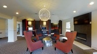 This single-floor space presents a thoughtfully designed interior that blends comfort with functionality. The room immediately draws attention with its dramatic spherical chandelier — a starburst fixture of clustered lights that serves as the centerpiece of the space, casting a warm, ambient glow across the room. The seating arrangement features high-backed wingback chairs upholstered in rich red fabric, paired with deep charcoal accent chairs, all gathered around a metallic gold side table, creating an inviting lounge area ideal for conversation or relaxation. The patterned carpet adds texture and warmth underfoot, complementing the bold color palette throughout. Along the perimeter, built-in dark cabinetry and workstation desks provide dedicated workspace, complete with computer monitors and task chairs, making this area equally suited for productivity. Accent lighting integrated into the cabinetry columns adds a layered lighting effect, enhancing the sophisticated ambiance. A window along one wall allows natural light to filter in, offering a counterbalance to the richly dark interior tones. The overall design aesthetic is contemporary and upscale, with a cohesive blend of bold colors, refined furnishings, and purposeful layout that serves both as a social gathering space and a functional business center.