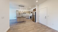 Located at 9860 W Tropicana Ave in Las Vegas, NV, this well-designed 2-bedroom, 1-bathroom residence offers a thoughtful single-floor layout with modern finishes throughout. The open-concept kitchen serves as the heart of the home, showcasing light wood cabinetry, a subway tile backsplash, dark quartz countertops, and a full suite of stainless steel appliances including a gas range, microwave, and refrigerator. A built-in wine rack adds a distinctive touch, while a central island with pendant lighting creates an ideal space for dining and entertaining. The kitchen flows seamlessly into the living area, which features wood-look flooring and a window that brings in natural light. Both bedrooms are carpeted and bright, with large windows offering outdoor views of the surrounding Las Vegas landscape. The primary bedroom is generously sized and includes a ceiling fan for added comfort. The bathroom is clean and contemporary, featuring wood-tone cabinetry, a backlit vanity mirror, and a full tub-shower combination with white tile surround. A dedicated closet provides practical storage. The overall aesthetic is fresh and modern, with a neutral palette of whites and warm wood tones creating a cohesive and inviting atmosphere throughout.