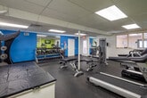 This single-floor amenity space is thoughtfully organized into distinct functional zones, each designed to serve residents efficiently. The fitness center is a well-appointed area featuring rubber flooring, drop-panel ceilings with recessed fluorescent lighting, and a wall-mounted flat-screen television. Equipment includes a treadmill, multi-function cable machine, weight benches, ab rollers, a medicine ball rack, and a padded exercise mat platform, providing a comprehensive workout environment. Full-length mirrors along one wall enhance the sense of openness and allow for proper form checks during exercise. Adjacent to the fitness area, a comfortable lounge zone provides a place to relax, furnished with cushioned armchairs and ottomans accented with striped pillows, a small wooden side table, and a bar-height counter with seating overlooking the fitness room through a pass-through window. The laundry facility is clean and efficiently arranged, featuring multiple stacked washer and dryer units with digital controls, durable tile flooring, and informational signage to guide users. A small trash receptacle is conveniently positioned nearby. Together, these spaces offer a practical and comfortable amenity suite catering to everyday resident needs.