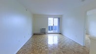 This well-maintained single-floor unit offers a clean, open layout bathed in natural light. The main living area is a standout feature, showcasing beautifully polished parquet hardwood flooring in a classic herringbone-style pattern that adds warmth and timeless character to the space. Crisp white walls throughout create a bright, airy ambiance that makes the room feel open and welcoming.

A large sliding glass door serves as the focal point of the living area, flooding the space with abundant natural light while providing direct access to a private balcony. The balcony overlooks a surrounding urban environment, offering open city views. A wall-mounted air conditioning unit and baseboard heating ensure year-round climate comfort.

The overall aesthetic of the space is clean and minimal, providing a neutral canvas well-suited for a variety of interior styles. The combination of gleaming parquet floors, generous natural light, and balcony access makes this a functional and inviting living space in an urban setting.