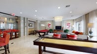 This single-floor amenity space on S Voss Rd in Houston, TX, is designed as a sophisticated social and recreational hub. The open layout is anchored by polished marble-style tile flooring that extends throughout, creating a seamless, upscale aesthetic. Recessed lighting illuminates the entire space, complemented by statement pendant fixtures that add visual interest overhead.

The game area is a central highlight, featuring a red felt billiard table with a distinctive curved base and a full-length shuffleboard table, both positioned on the open floor. A lounge zone nearby offers plush seating, including bold red accent chairs, a tufted ottoman, and a sofa arrangement centered around a flat-screen television mounted on a dark granite media console with rich wood cabinetry.

The kitchen area is well-appointed with dark granite countertops, warm wood cabinetry, stainless steel appliances including a refrigerator and oven, and a bar-height island with red upholstered barstools. Vibrant abstract artwork adorns the walls throughout, reinforcing the contemporary design. Floor-to-ceiling windows along one wall flood the space with natural light and provide direct views of an outdoor pool courtyard, connecting the interior to the surrounding environment.