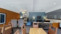 This well-appointed single-floor lounge space combines contemporary design with a warm, inviting ambiance. The interior is defined by a striking color palette, featuring bold teal and burnt orange accent walls that create distinct visual zones throughout the open layout. Recessed ceiling lighting is complemented by decorative pendant and table lamps, ensuring a well-lit yet comfortable atmosphere throughout the day and evening. The space is thoughtfully arranged with multiple seating areas to accommodate a variety of uses. A dining area anchors one end of the room with a sleek wood-top table surrounded by upholstered chairs, while the lounge area offers a mix of patterned high-back chairs, a plush sofa, and accent seating. A fireplace with a classic mantle serves as a focal point in one section of the room, adding a touch of elegance. On the opposite side, a mounted flat-screen television and media console provide entertainment options. Decorative artwork and floral arrangements add refined finishing touches throughout. The hardwood-style flooring ties the entire space together, contributing to a clean, modern aesthetic that balances functionality with style.