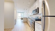 This single-floor unit presents a clean, contemporary design highlighted by light hardwood flooring that runs seamlessly through every room, creating a cohesive and airy atmosphere. The galley-style kitchen is well-appointed with white shaker cabinetry extending to the ceiling, stainless steel appliances including a range, microwave, dishwasher, and refrigerator, along with a neutral-toned countertop and a stainless steel sink. Track lighting overhead complements pendant lights that hang above the peninsula, which provides additional workspace and casual seating. The kitchen flows naturally into the living area, where large floor-to-ceiling windows flood the space with natural light and offer open views of the surrounding cityscape. The two bedrooms are calm and understated, each featuring the same light hardwood flooring, neutral walls, and ample closet access. The bathroom is finished with white cabinetry, a white countertop vanity with a large mirror, and a full shower-tub combination with white tile surround. The entryway provides a practical transition into the unit, with a closet nearby for storage. Throughout, the design emphasizes a bright, minimal aesthetic with a consistent neutral palette.