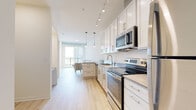 This single-floor unit presents a clean, contemporary design highlighted by light hardwood flooring that runs seamlessly through every room, creating a cohesive and airy atmosphere. The galley-style kitchen is well-appointed with white shaker cabinetry extending to the ceiling, stainless steel appliances including a range, microwave, dishwasher, and refrigerator, along with a neutral-toned countertop and a stainless steel sink. Track lighting overhead complements pendant lights that hang above the peninsula, which provides additional workspace and casual seating. The kitchen flows naturally into the living area, where large floor-to-ceiling windows flood the space with natural light and offer open views of the surrounding cityscape. The two bedrooms are calm and understated, each featuring the same light hardwood flooring, neutral walls, and ample closet access. The bathroom is finished with white cabinetry, a white countertop vanity with a large mirror, and a full shower-tub combination with white tile surround. The entryway provides a practical transition into the unit, with a closet nearby for storage. Throughout, the design emphasizes a bright, minimal aesthetic with a consistent neutral palette.
