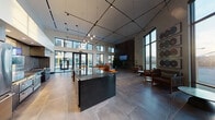 Located at 1101 N Main St in Los Angeles, CA, this single-floor amenity space offers a striking blend of modern industrial design and sophisticated comfort. The open-plan layout is anchored by soaring double-height ceilings, large-format stone tile flooring, and an abundance of natural light streaming through floor-to-ceiling windows that frame views of the surrounding urban landscape and courtyard pool area.

At the heart of the space is a fully appointed communal kitchen, featuring a dramatic dark stone island with bar seating, a gas cooktop with a professional hood, stainless steel refrigerator, tile backsplash, and sleek dark cabinetry. Industrial-style pendant track lighting overhead adds warmth and visual interest to the kitchen zone.

Adjacent to the kitchen, a spacious lounge area invites relaxation with a large sectional sofa, layered rugs, and curated coffee tables — all positioned to take full advantage of the panoramic window views. A separate seating nook near the perimeter windows features leather armchairs, bistro tables, and a series of vibrant circular artwork panels, creating an intimate yet airy atmosphere.

A wall-mounted display screen and a stone-clad accent wall add functional and aesthetic dimension to the space, while lush indoor plants contribute a natural, welcoming energy throughout.