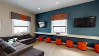 Located at 435 Reflections Circle in San Ramon, California, this single-floor community lounge space is designed with a modern, energetic aesthetic that makes it well-suited for both relaxation and casual collaboration. The interior is defined by a striking color palette — bold teal accent walls paired with vibrant orange furnishings create a lively, welcoming atmosphere throughout.

The main lounge area features a large, comfortable gray sectional sofa and ottoman, providing ample seating for gatherings. A row of round orange ottomans lines the teal feature wall, beneath a continuous built-in white shelf that serves as a convenient workspace or surface for personal items. Multiple flat-screen TVs are mounted on the walls, offering entertainment from various vantage points throughout the room.

Natural light enters through several windows dressed with coordinating brown and orange Roman shades, keeping the space bright and inviting. The light-toned carpet tile flooring adds warmth and texture underfoot.

A dedicated nook provides a more intimate seating arrangement, complete with a round dining table, retro-style wire chairs, and colorful accent pillows. Additional functional amenities include a multifunction printer and storage closets, making this lounge as practical as it is stylish. Decorative touches such as a circular wall mirror and small potted plants add personality and a finishing polish to the overall design.