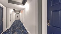 This single-floor corridor space presents a refined and polished interior design that balances both functionality and aesthetic appeal. The hallway immediately draws attention with its bold, navy blue and gold marbled patterned carpeting, which runs the full length of the passage and serves as a dramatic visual centerpiece against the clean, white walls and bright white baseboards.

The ceiling design adds architectural interest through the use of structured coffered detailing with dark gray accent beams, lending a sense of depth and dimension overhead. Wall-mounted sconces are evenly distributed along the corridor, providing warm, consistent ambient lighting that enhances the sophisticated atmosphere.

The walls are finished in a crisp, neutral white, which contrasts elegantly with the darker tones of the ceiling and flooring. A framed piece of abstract artwork is displayed at the far end of the hallway, serving as a focal point that draws the eye down the length of the space. Dark navy doors with polished hardware line the corridor, adding further contrast and a sense of intentional color coordination throughout.

Emergency signage is discreetly integrated into the design without detracting from the overall aesthetic. The overall ambiance of the space is one of modern sophistication, with careful attention to detail in every design element, from the layered lighting to the cohesive color palette.