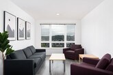 This well-appointed multi-bedroom residence is designed with a clean, modern aesthetic throughout. The open-plan living area is anchored by large windows that frame sweeping city and treetop views, flooding the space with natural light. Light wood-tone flooring runs consistently throughout, lending a cohesive and airy feel. The living room comfortably accommodates a sofa and armchairs, with wall space featuring contemporary framed artwork that adds a subtle decorative touch.

The kitchen is fully equipped for everyday use, featuring light wood cabinetry extending to the ceiling, dark countertops, a breakfast bar peninsula, and full black appliances including a range, microwave, and dishwasher. Recessed lighting ensures the kitchen remains bright and functional.

Each private bedroom is thoughtfully furnished with a single bed frame, a dedicated study desk and ergonomic chair, and a window providing natural light and outdoor views — making them well-suited for focused work or rest. The clean white walls and light flooring create a calm, neutral environment in every room.

The bathroom is bright and practical, featuring a shower-tub combination with a curtain, toilet, and vanity with storage, all finished in a consistent light palette with wood-look flooring. The unit's entry opens directly into the kitchen and living areas, offering a straightforward and efficient layout.