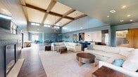 Located at 2400 Buckner Ln in Spring Hill, Tennessee, this generously sized single-floor space offers a thoughtfully designed open-concept layout that seamlessly connects the kitchen, living room, and dining areas. The great room commands immediate attention with its dramatically high ceilings adorned with exposed wood beams and a striking chandelier, while wood-look tile flooring and blue-toned shiplap walls establish a cohesive, modern-farmhouse aesthetic throughout. The living area is anchored by plush white sofas, oval bronze accent tables, and an area rug, creating an inviting gathering space bathed in natural light from large windows overlooking the outdoor courtyard. The kitchen is a standout feature, showcasing a large waterfall-edge quartz island with bar seating, dark espresso cabinetry, stainless steel appliances, white countertops, and pendant lighting — all designed for both functionality and style. A dedicated seating nook with leather chairs and oversized windows provides a quieter retreat within the open floor plan. The formal dining room is equally impressive, featuring a tray ceiling with a cage-style pendant light, a long solid wood dining table with upholstered seating, blue wainscoting, textured upper walls, and framed artwork — making it ideal for gatherings and events.