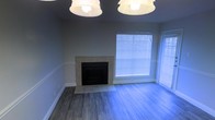 This single-floor space presents a clean, understated living area designed with functional simplicity in mind. The focal point of the room is a tile-surround fireplace, framed in neutral-toned tiles that extend to a modest hearth, adding both warmth and architectural character to the space. The fireplace is set against cool gray walls, contributing to a calm, contemporary aesthetic throughout.

The flooring consists of hardwood-style planks in a soft gray tone, lending a modern yet inviting feel underfoot. Two pendant light fixtures are mounted overhead, providing even illumination across the room and complementing the overall neutral color palette.

Natural light enters through large windows fitted with white horizontal blinds, allowing for adjustable brightness while maintaining privacy. The windows add a sense of openness to the room, enhancing the airy quality of the space.

White baseboards provide a crisp contrast against the gray walls and flooring, reinforcing the clean lines found throughout. Overall, the space presents a well-lit, move-in-ready environment with a cohesive, neutral design scheme suitable for a variety of furnishing styles.