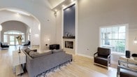 This contemporary living space showcases an open floor plan that seamlessly blends various functional areas. The room is characterized by soaring ceilings that create a sense of spaciousness and grandeur. A focal point of the space is the sleek fireplace, framed by a tall, navy blue accent wall that adds depth and visual interest.

The living area features comfortable seating, including a plush gray sofa and modern armchairs, arranged atop a geometric patterned rug. Adjacent to this, a stylish kitchen boasts dark wood cabinetry, stainless steel appliances, and a convenient breakfast bar with modern stools.

Large windows and glass doors flood the space with natural light, while strategically placed recessed lighting and wall sconces provide additional illumination. The hardwood floors throughout contribute to the cohesive, refined aesthetic.

An arched entryway leads to another seating area, where tufted armchairs and a unique circular coffee table create an inviting nook. The room's neutral color palette, primarily whites and grays, is punctuated by carefully chosen decor elements, fostering a sophisticated and serene atmosphere.