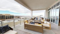 This rooftop terrace is a thoughtfully designed outdoor living space that seamlessly blends covered and open-air zones for a versatile and inviting atmosphere. The terrace is finished with wood-look porcelain tile flooring throughout, providing a clean, contemporary foundation that complements the modern architectural design.

The covered portion of the terrace features floor-to-ceiling sliding glass doors and expansive transom windows that connect the outdoor space to the interior, allowing natural light to flow freely between the two. Decorative patterned window film adds a refined, geometric design element to the glazing. Recessed ceiling lighting and structural columns define this sheltered seating area, which accommodates multiple conversation groupings of teak-framed and wicker lounge furniture upholstered in crisp white with bold navy, yellow, and coral accent pillows.

Beyond the covered zone, the open-air terrace extends generously, offering additional seating arrangements anchored by striped area rugs and low teak coffee tables. Glass panel railings line the perimeter, preserving unobstructed panoramic views of the surrounding cityscape, rolling hills, and distant mountain ranges. A built-in daybed tucked against a structural column provides an intimate nook ideal for relaxing and taking in the sweeping skyline. The overall ambiance is bright, modern, and resort-inspired.