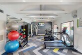 Located at 9940 Richmond Ave in Houston, TX, this fitness center offers a comprehensive workout environment designed to support a wide range of exercise routines. The space is defined by its vaulted ceiling with prominent exposed white beams, lending an open and airy architectural character throughout.

The flooring features a bold chevron-patterned carpet in shades of gray, blue, and yellow, adding a dynamic visual element that complements the modern, energetic atmosphere. A large wall-mounted flat-screen TV is positioned near a deep gray accent wall, providing entertainment during workouts.

Cardio equipment includes TRUE-brand treadmills and elliptical machines, as well as dedicated cycling bikes stationed near a large picture window that frames a direct view of the outdoor pool area. This window floods the space with natural light and connects the interior to the surrounding amenity areas. A multi-station cable machine supports strength training, while a well-stocked rack of medicine balls, stability balls, and kettlebells offers versatile free-weight options.

Full-length mirrors along one wall visually expand the space and assist with form during exercise. The fitness center is accessed through elegant double glass doors and includes a sanitizing station, fire safety equipment, and informational signage. Natural light also enters through additional windows, keeping the environment bright and inviting throughout the day.