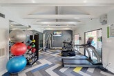Located at 9940 Richmond Ave in Houston, TX, this fitness center offers a comprehensive workout environment designed to support a wide range of exercise routines. The space is defined by its vaulted ceiling with prominent exposed white beams, lending an open and airy architectural character throughout.

The flooring features a bold chevron-patterned carpet in shades of gray, blue, and yellow, adding a dynamic visual element that complements the modern, energetic atmosphere. A large wall-mounted flat-screen TV is positioned near a deep gray accent wall, providing entertainment during workouts.

Cardio equipment includes TRUE-brand treadmills and elliptical machines, as well as dedicated cycling bikes stationed near a large picture window that frames a direct view of the outdoor pool area. This window floods the space with natural light and connects the interior to the surrounding amenity areas. A multi-station cable machine supports strength training, while a well-stocked rack of medicine balls, stability balls, and kettlebells offers versatile free-weight options.

Full-length mirrors along one wall visually expand the space and assist with form during exercise. The fitness center is accessed through elegant double glass doors and includes a sanitizing station, fire safety equipment, and informational signage. Natural light also enters through additional windows, keeping the environment bright and inviting throughout the day.