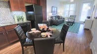 This single-floor residence showcases an open-concept layout that seamlessly connects the kitchen, dining, and living areas into one cohesive and functional space. The kitchen is outfitted with rich, dark wood cabinetry, granite countertops, and black appliances, including a full-size refrigerator and dishwasher, lending a modern and polished aesthetic. A tiled backsplash adds a subtle decorative touch to the kitchen area.

Adjacent to the kitchen, the dining area accommodates a round dark table paired with sleek black leather chairs, making it ideal for casual meals. The hardwood-style flooring runs throughout, providing a warm and unified foundation across the living spaces.

The living room extends naturally from the dining area, featuring large windows that allow generous natural light to fill the space. A patterned area rug anchors the seating arrangement, which includes a dark sofa and an accent chair in a bold chevron fabric. The overall ambiance is contemporary and inviting, with a neutral color palette accented by vibrant décor elements.