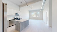 This contemporary single-floor unit showcases a thoughtfully designed open-concept layout that seamlessly integrates the kitchen, dining, and living areas. Soaring ceilings with exposed industrial-style ductwork and track lighting lend the space a loft-like character, creating an airy and expansive atmosphere throughout.

The kitchen is a standout feature, equipped with stainless steel appliances including a refrigerator, range, and built-in microwave. Crisp white upper cabinetry pairs elegantly with warm, wood-toned lower cabinets and a matching kitchen island topped with a clean white surface — offering both additional prep space and a natural gathering point. A white subway tile backsplash completes the polished look.

Light hardwood-style flooring flows continuously throughout the main living areas, enhancing the sense of openness. Large windows in the living area and bedroom flood the space with natural light and provide views of the surrounding neighborhood. The bedroom is generously proportioned with high ceilings and a large multi-pane window that further brightens the room.

The bathroom features elegant marble-look tile flooring, a wood-toned vanity with a white countertop, and brushed nickel fixtures, delivering a refined and cohesive aesthetic. Overall, the unit presents a clean, modern design with industrial accents and a bright, welcoming ambiance.