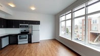 This single-floor space offers a clean, modern aesthetic defined by an open-concept layout that seamlessly connects the kitchen and living area. The kitchen is outfitted with dark cabinetry, stainless steel appliances — including a range, microwave, and refrigerator — and is thoughtfully designed to maximize functionality within the space. Warm hardwood-style flooring runs throughout, adding a welcoming contrast to the crisp white walls and clean lines of the interior. A standout feature of the living area is the expansive multi-pane window that floods the space with natural light, creating an airy and open atmosphere. The oversized window also frames views of the surrounding neighborhood, enhancing the sense of connection to the outdoors. The neutral color palette, smooth ceilings, and minimalist design make this space versatile and easy to personalize. Overall, the unit strikes a balance between practicality and style, offering a comfortable and well-lit environment suited to a variety of living arrangements.