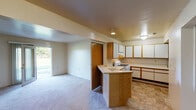 This single-story residence offers a practical and functional layout distributed across several interconnected rooms. The kitchen serves as a central hub of the home, featuring an abundance of white upper cabinetry with wood trim accents, generous counter space, a gas range, and a sink positioned on a peninsula that opens toward the living area. The tile flooring in the kitchen provides a durable, easy-to-maintain surface. A built-in peninsula with warm wood paneling visually connects the kitchen to the adjacent living space, which is finished with wall-to-wall carpeting and neutral-toned walls throughout. The living area benefits from a sliding glass door that allows natural light to filter in while providing direct access to the outdoors. Additional rooms, also carpeted, offer versatile spaces with windows that bring in natural light and views of surrounding greenery. The full bathroom includes a tub-shower combination with a surround, a single-sink vanity with wood cabinetry, a large mirror, and tile flooring. Overall, the residence presents a straightforward, well-organized floor plan with consistent neutral finishes and practical amenities throughout.